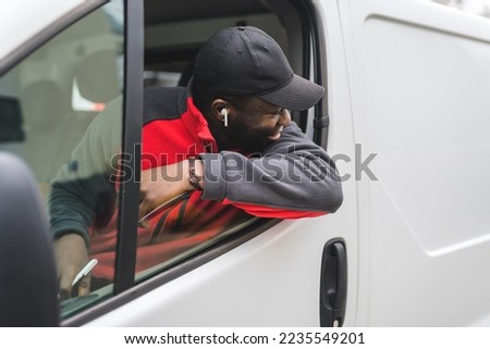 Black young adult delivery man sitting in driver seat of white van leaning out the window smiling looking away from camera. Horizontal shot. High quality photo