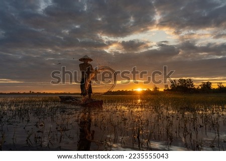 Asian village lifestyle concept. A male asian mature adult fisherman cast fishing net in a nearby freshwater lake during dawn sunrise in Sakon Nakhon, Northeast Thailand, Isan way of life. Royalty-Free Stock Photo #2235555043