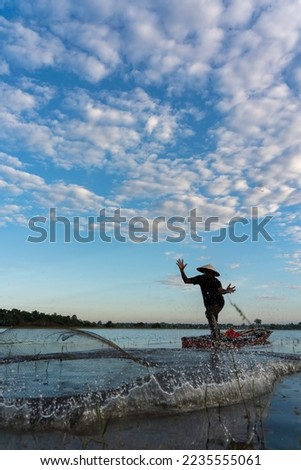 Asian village lifestyle concept. A male asian mature adult fisherman cast fishing net in a nearby freshwater lake during dawn sunrise in Sakon Nakhon, Northeast Thailand, Isan way of life. Royalty-Free Stock Photo #2235555061