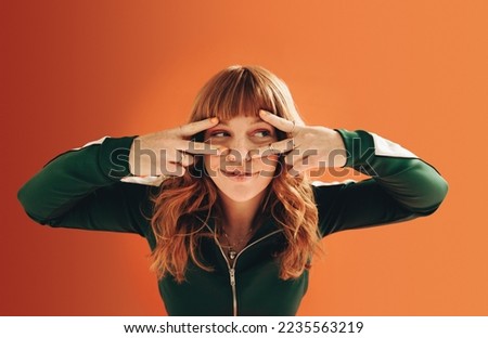 Woman with ginger hair making v signs with her hands. Young woman having fun against an orange background. Royalty-Free Stock Photo #2235563219