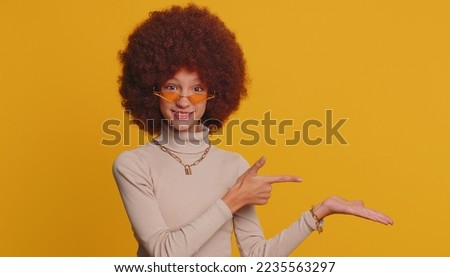 Happy funny girl showing thumb up pointing empty place, advertising area for commercial text, copy space for goods promotion. Young little child kid. Studio shot indoors on yellow wall background