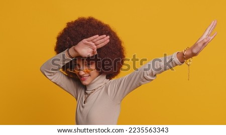 Trendy cheerful positive young school girl with afro hairstyle having fun dancing and moving to rhythm, dabbing raising hands, making dubdance gesture. Teenager female child kid on yellow background Royalty-Free Stock Photo #2235563343
