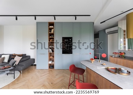 Interior design of colorful open space with built-in oven, bookcase, modern red hockers, wooden kitchen island, gray sofa, pillows, panels floor and personal accessories. Home decor. Template. Royalty-Free Stock Photo #2235569769