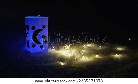 Photography of a Chinese paper lantern. Dark winter night with moon and stars are drawing on it. White snow  as background. Golden lighs of flashlight on the snow. 