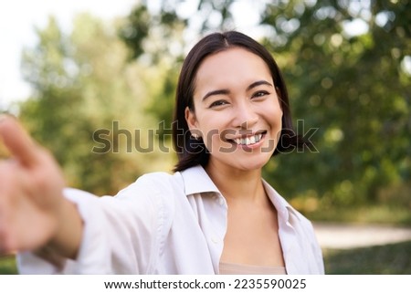 People and lifestyle. Happy asian woman takes selfie in park, photo on smartphone, smiling and looking joyful.