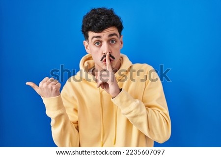 Hispanic man standing over blue background asking to be quiet with finger on lips pointing with hand to the side. silence and secret concept. 