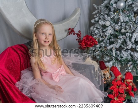 A little blonde girl in a pink dress next to a Christmas tree on the background of a decorative moon. Magical Christmas time.
