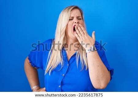 Caucasian plus size woman standing over blue background bored yawning tired covering mouth with hand. restless and sleepiness. 