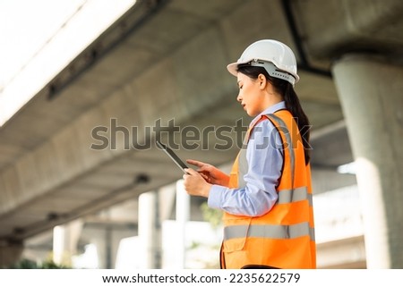 Asian engineer worker woman or architect looking construction with tablet wear white safety helmet in construction site. Standing at highway concrete road site. Progress planning of highway bridge.