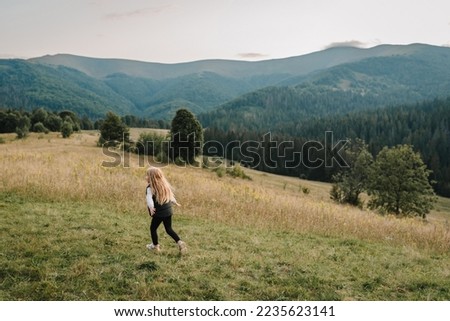 Child in autumn mountains. Girl is running on the grass in the field and enjoying nature. Little hiker and traveler walk on top of foggy mountain. World Tourism Day. Active outdoor games. Back view.