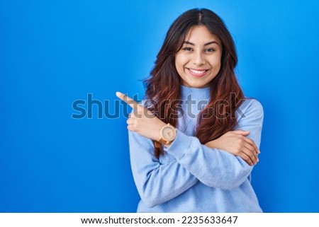 Hispanic young woman standing over blue background with a big smile on face, pointing with hand finger to the side looking at the camera. 