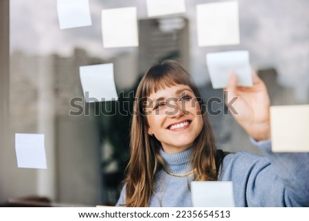 Cheerful young businesswoman sticking adhesive notes to a glass wall in a modern office. Happy female entrepreneur laying out her ideas while working on a new creative strategy. Royalty-Free Stock Photo #2235654513