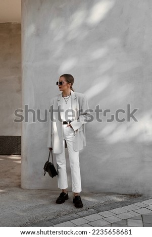 beautiful girl dressed in white flared jeans, gray jacket, t-shirt, black patent leather loafers, bag, sunglasses, belt, hair gathered in tail, accessories, stylish fashion outfit, lifestyle model Royalty-Free Stock Photo #2235658681