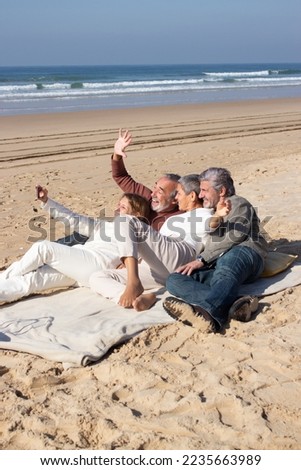 Cheerful senior friends taking selfie at seashore on sunny day. Two happy couples having fun while woman in white clothes holding smartphone and taking photo. Leisure, modern technology concept