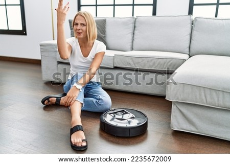 Young caucasian woman sitting at home by vacuum robot doing italian gesture with hand and fingers confident expression 