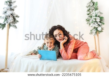 Family Christmas morning. Beautiful family mother and little daughter in pajamas watching movie or cartoon together on modern digital tablet while relaxing on canopy bed decorated with xmas lights