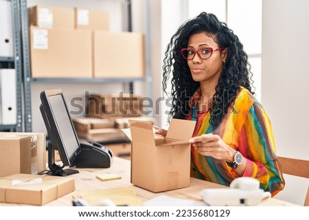 Middle age hispanic woman working at small business ecommerce looking inside box smiling looking to the side and staring away thinking. 