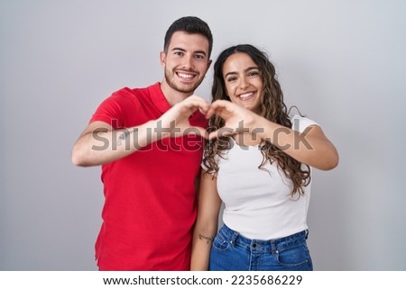 Young hispanic couple standing over isolated background smiling in love doing heart symbol shape with hands. romantic concept. 