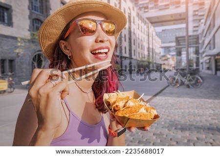 Happy cheerful girl eating spring rolls from takeaway paper box using chopsticks at Cologne city street. Fast food eatery and asian cuisine Royalty-Free Stock Photo #2235688017
