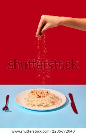 Female hand salting plate with pearl necklaces symbolizing noodles, pasta on blue tablecloth over red background. Food pop art photography. Complementary colors. Copy space for ad, text