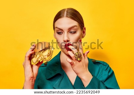 Young beautiful woman in green coat with red lipstick eating cheeseburger with necklaces, licking fingers over yellow background. Food pop art photography. Complementary colors. Copy space for ad