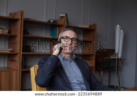 Portrait of elderly worried grey haired businessman in blue suit and eye glasses talking on mobile phone on grey studio background with bookshelves
