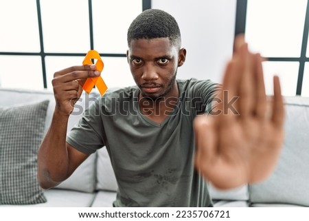 Young african american man holding awareness orange ribbon sitting on the sofa with open hand doing stop sign with serious and confident expression, defense gesture 