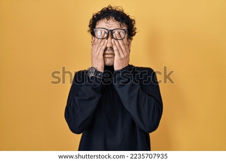 Hispanic man standing over yellow background rubbing eyes for fatigue and headache, sleepy and tired expression. vision problem 