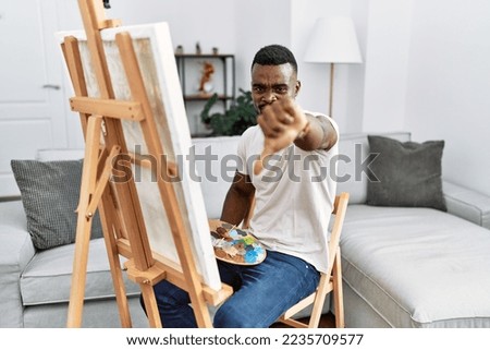 Young african man painting on canvas at home looking unhappy and angry showing rejection and negative with thumbs down gesture. bad expression. 