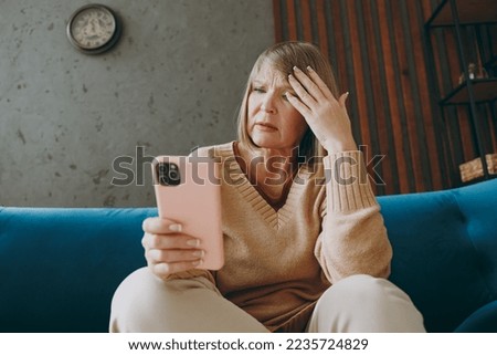 Elderly sad woman 50s year old wear casual clothes sit on blue sofa hold do not know how to use mobile cell phone prop up head stay at home flat spend free spare time in living room indoor grey wall Royalty-Free Stock Photo #2235724829