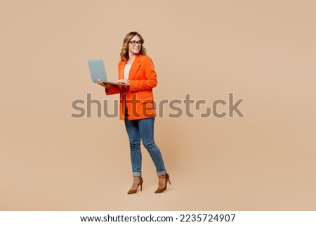 Full body fun young employee IT business woman corporate lawyer wear classic formal orange suit glasses work in office hold use laptop pc computer look aside on area isolated on plain beige background