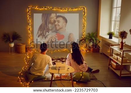 Romantic couple having Valentine's Day date at home, using modern projector in living room interior with beautiful fairy lights, watching movie, looking at digital photos, enjoying memories. Back view