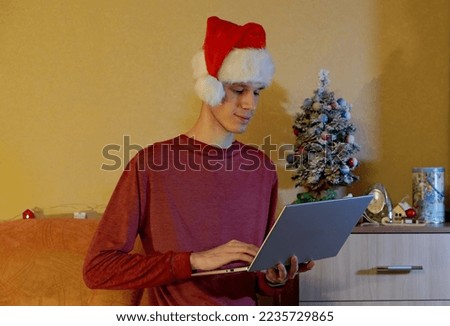 a young man in a Santa Claus hat buys gifts on a laptop online against the background of a bedside table with a small Christmas tree and gifts in the room