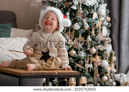 A beautiful little boy in a Santa Claus hat is sitting on a table and laughing merrily against the background of a Christmas tree and beautiful bokeh. The concept of celebrating Christmas.