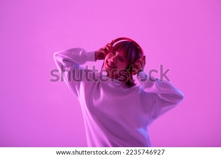 Young Asian woman wearing wireless headphones listening music in neon light background. Happy motions and facial expression concept	
