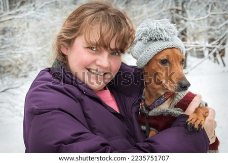 In winter, in a snowy forest, on a frosty day, a girl holds a dachshund dog in her arms in a hat and clothes. 