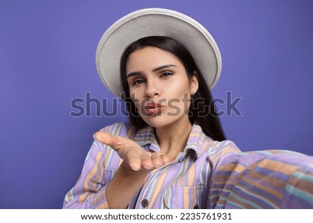 Beautiful young woman with stylish hat taking selfie while blowing kiss on purple background, closeup
