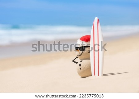 Sandy Christmas Snowman is watching the waves, standing on beautiful beach with a surf board, wearing sunnies