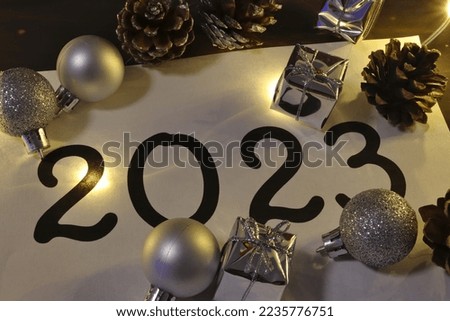 illuminated black inscription 2023 on a white background on which silver ornaments and cones are freely distributed