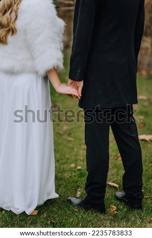 The groom in a black coat and the bride in a white fur coat are walking in the park with green grass on their backs, holding hands in autumn. Wedding photography, portrait.