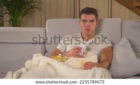 CLOSE UP: Young man eating snacks and watching latest television news report. Handsome guy covered with warm blanket getting upset while following TV news. Man on a comfy sofa watching TV report.