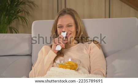 CLOSE UP: Lady smiling while watching entertainment TV program on a comfy sofa. Young woman covered with blanket and eating snacks while watching movie. Winter evenings in comfort of home living room.