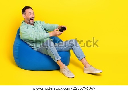 Full length photo of impressed funky guy dressed green hoodie sitting bean bag playstation empty space isolated yellow color background