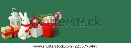 Banner on a green background, a white hare is a symbol of the year, a red cup with marshmelo and New Year's ice cubes.  Christmas and New Year concept, festive atmosphere.  Space for copy text.