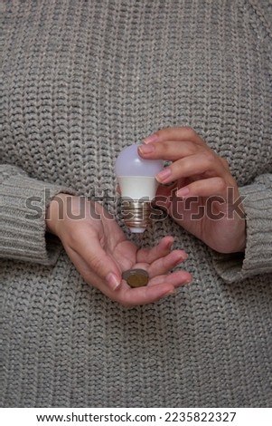 Woman in a gray knitted sweater holds a light bulb and coins in her palm in female hands. The concept of payment for energy, debt for electricity. Raising the price of utility bills. Financial crisis Royalty-Free Stock Photo #2235822327