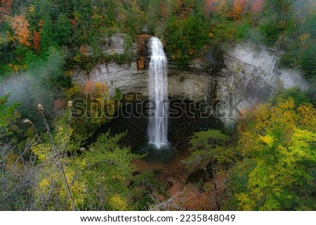 Falls Creek Falls is surrounded by vibrant fall colors and a misty morning haze. The waterfall cascades from a rocky cliff into a dark pool below. Photographed  at Falls Creek Falls State Park in TN.  Royalty-Free Stock Photo #2235848049