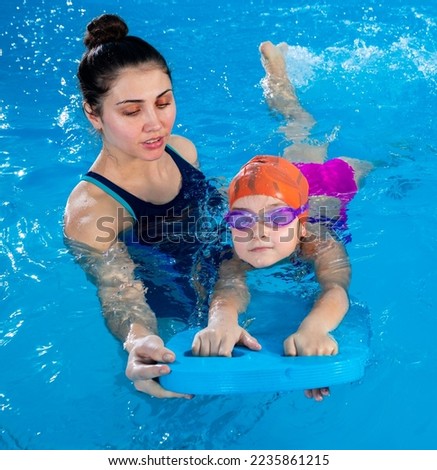 Trainer teaching little girl how to swim in indoor pool with pool floating board