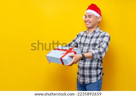 Excited young Asian man wearing plaid shirt with a Santa Claus hat giving a gift box isolated on yellow studio background. Merry Christmas Concept