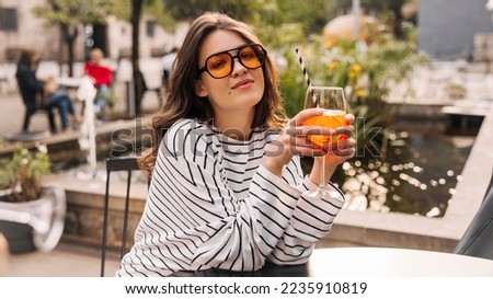 Pretty caucasian young lady is enjoying cooling drink sitting in cafe on hot day. Brunette wears sunglasses, light clothes. Relaxation concept