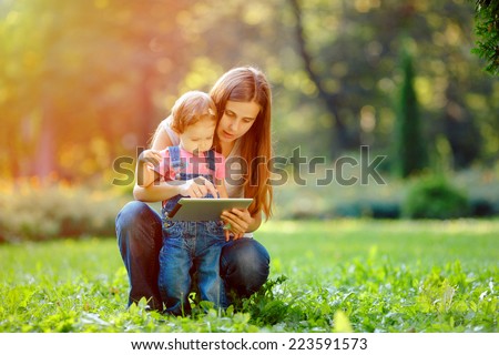 Beautiful mother and kid girl playing at tablet pc outdoors in fall. Happy mother showing her daughter funny pictures your tablet. Happy family, mother and daughter with tablet resting in the park.